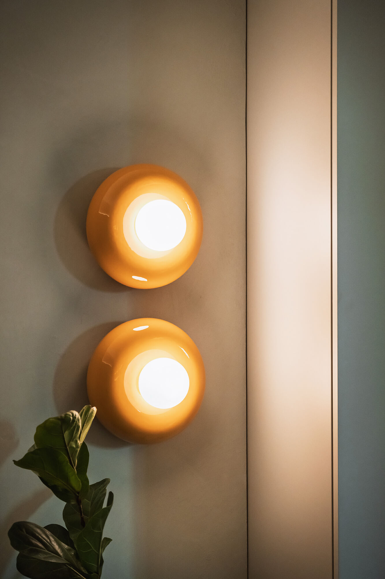 Dwa pomarańczowe kinkiety na ścianie, obok roślina doniczkowa. Nowoczesne oświetlenie.