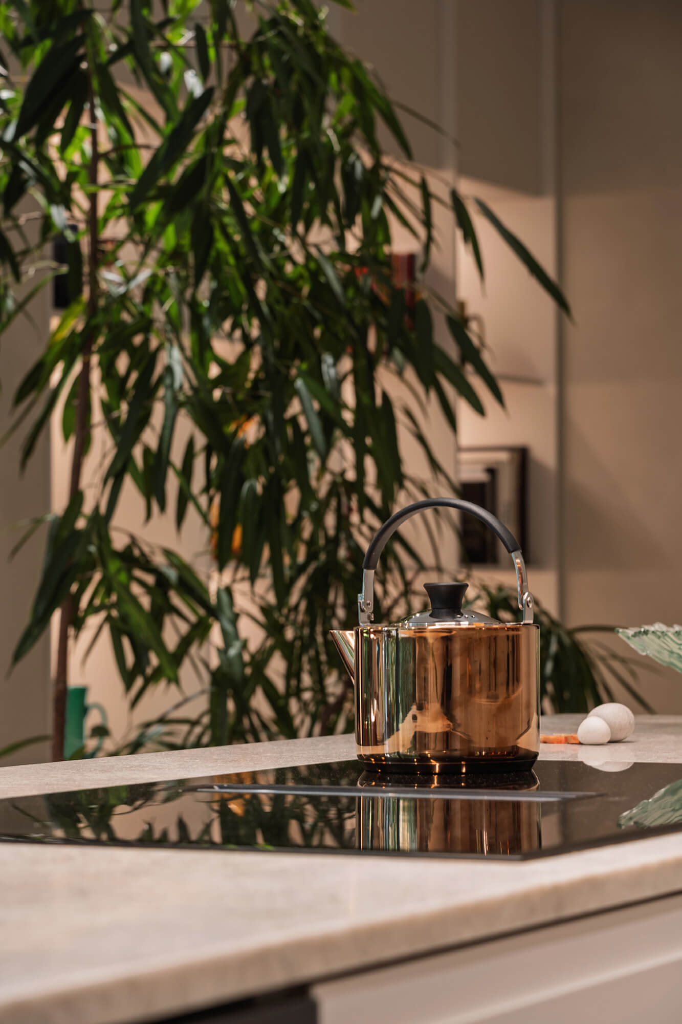 Miedziany czajnik na płycie indukcyjnej w nowoczesnej kuchni z rośliną w tle.
