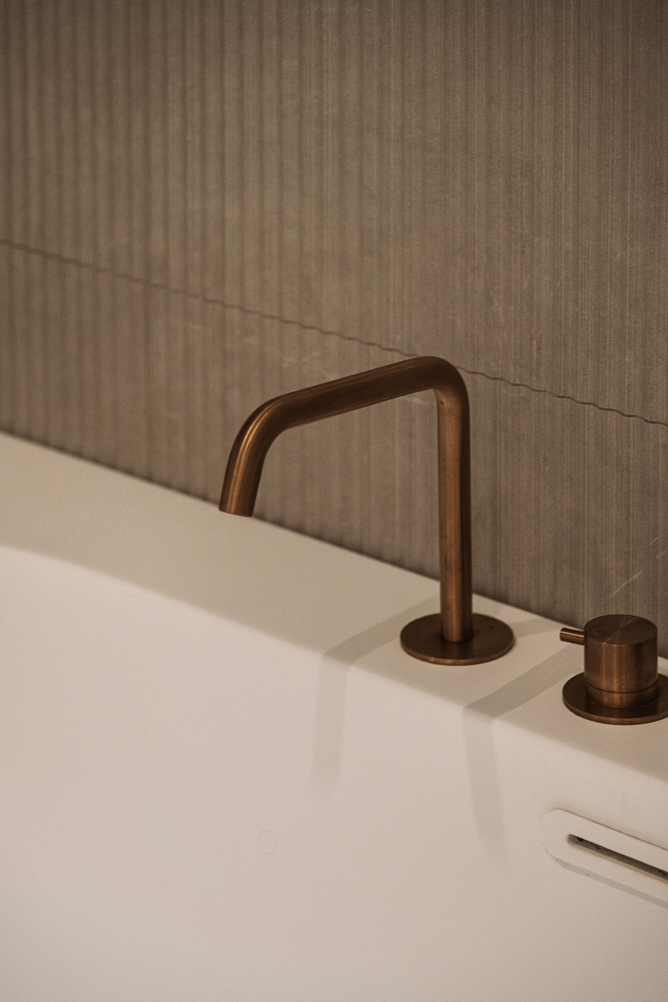 Miedziana bateria umywalkowa na białej wannie, na tle szarych płytek.