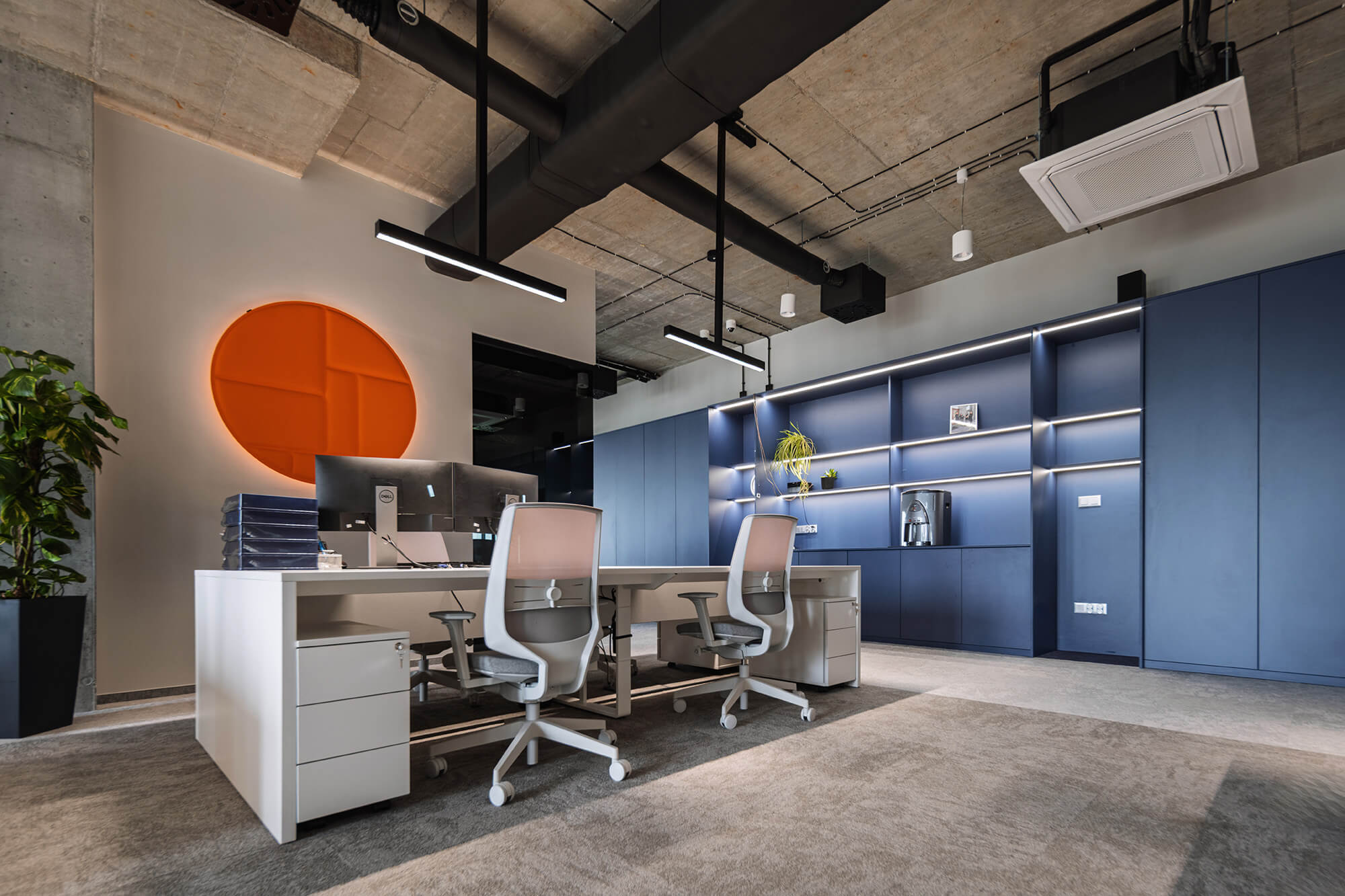 Nowoczesne biuro z betonowym sufitem, niebieską zabudową i pomarańczowym kołem na ścianie.