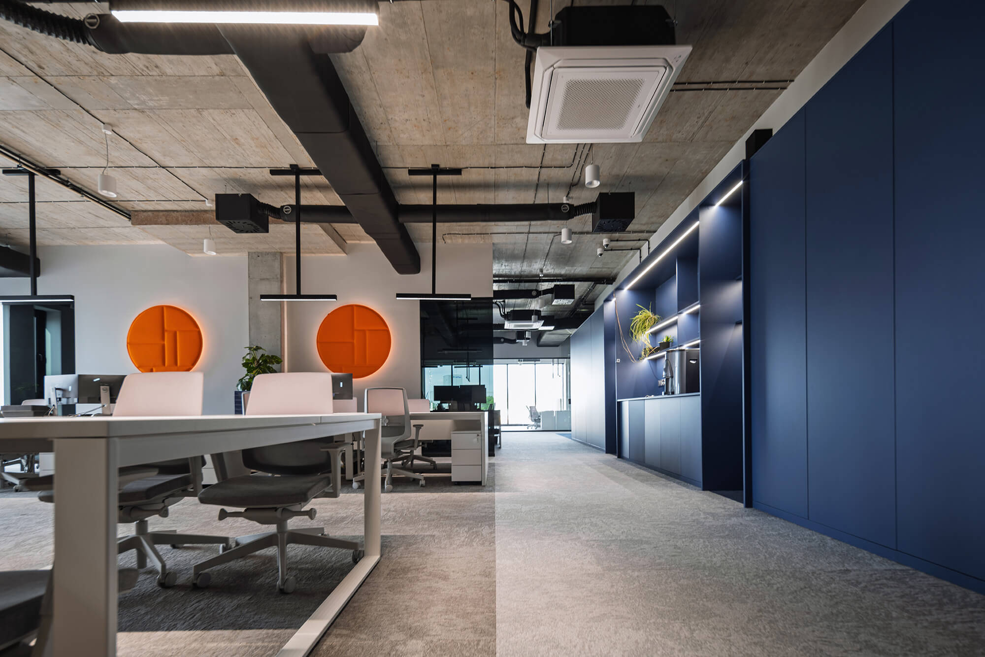 Nowoczesne biuro z otwartą przestrzenią, szare biurka, pomarańczowe dekoracje i surowy betonowy sufit.
