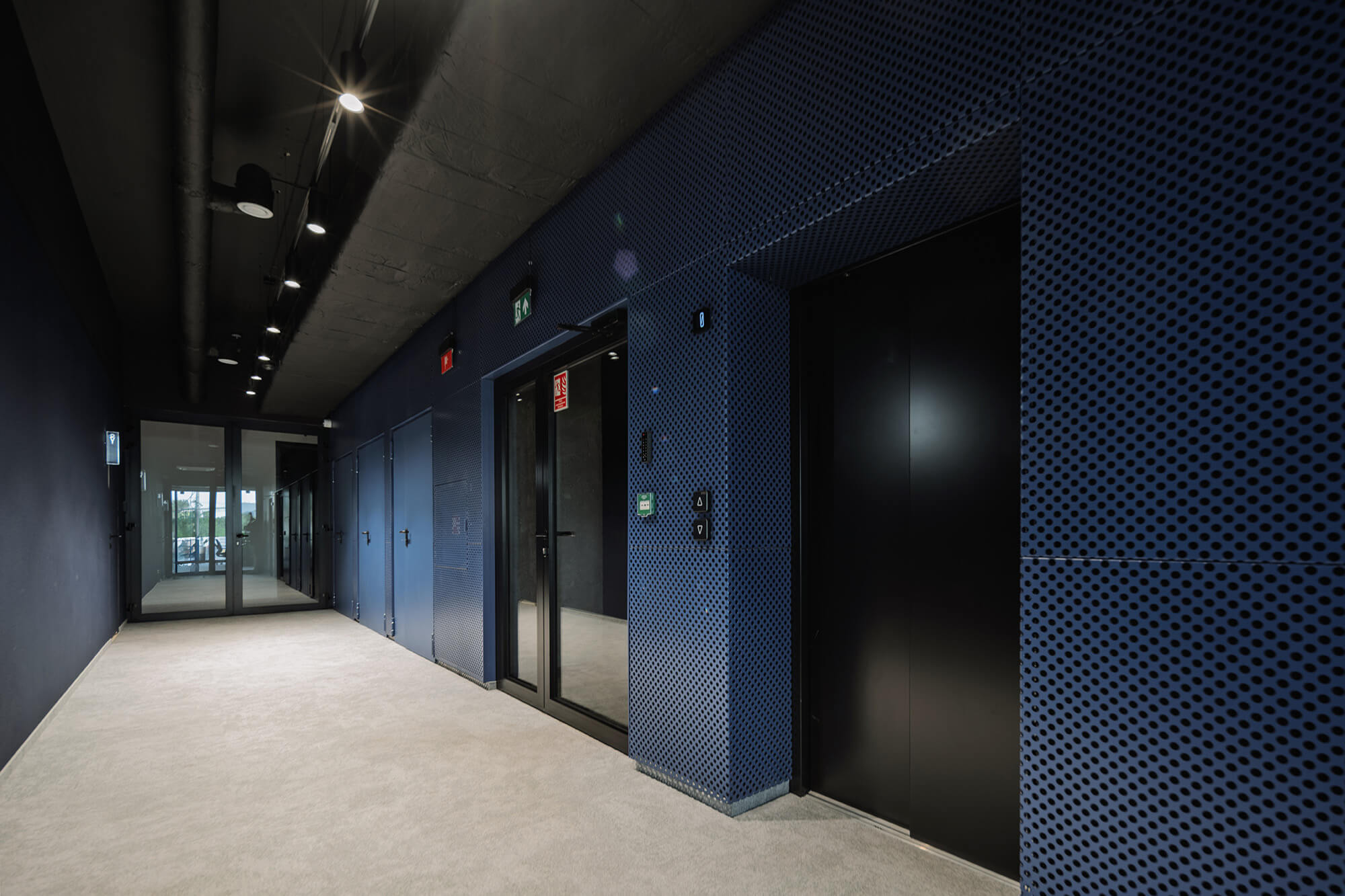 Korytarz z niebieskimi panelami perforowanymi, czarnymi drzwiami i jasną podłogą.