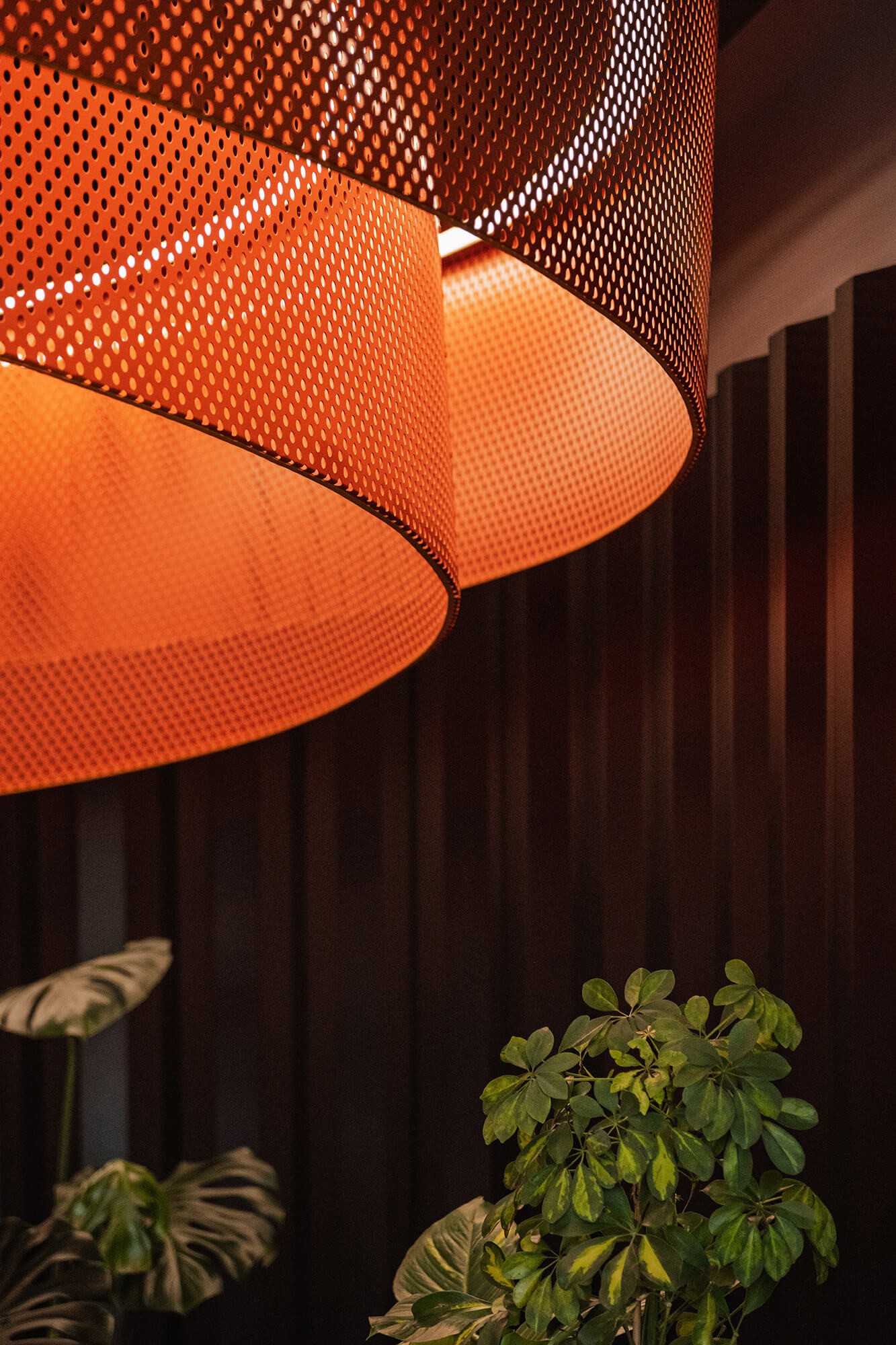 Pomarańczowa lampa z perforowanego metalu i zielone rośliny na tle ściany.