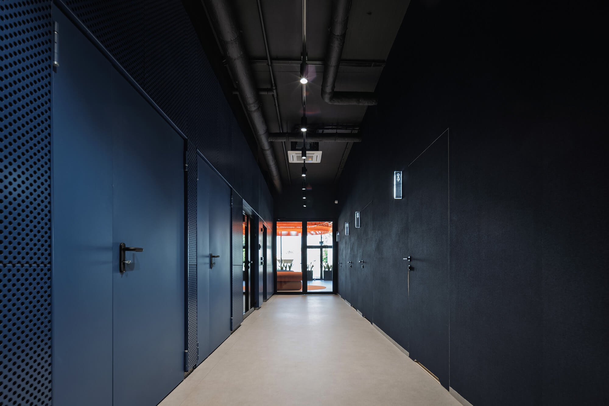 Korytarz z niebieskimi drzwiami i czarnymi ścianami prowadzący do jasnego pomieszczenia.