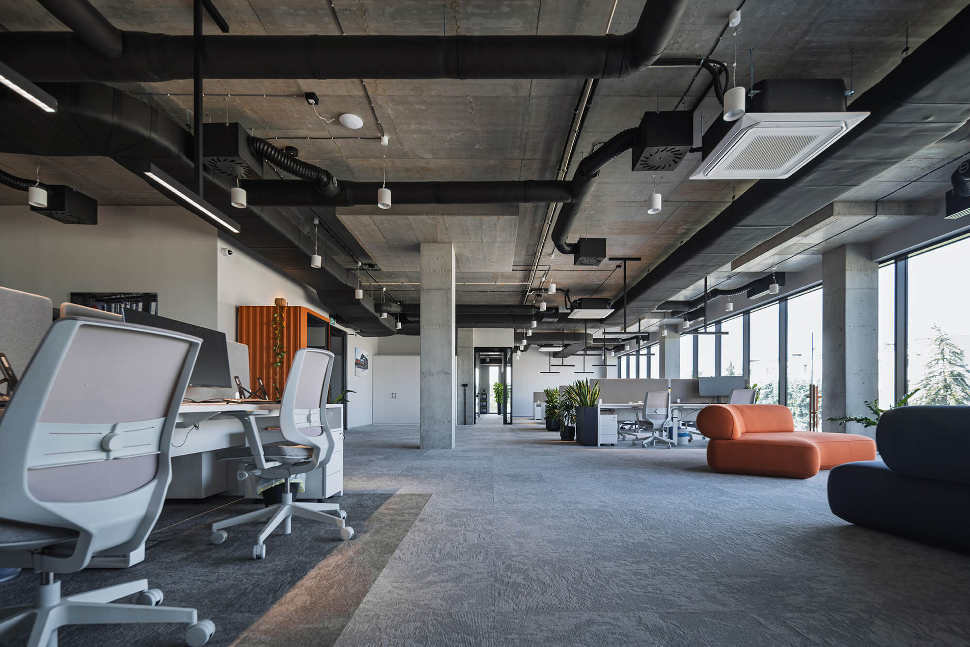 Nowoczesne biuro z otwartą przestrzenią, szary dywan i betonowy sufit z czarnymi rurami.