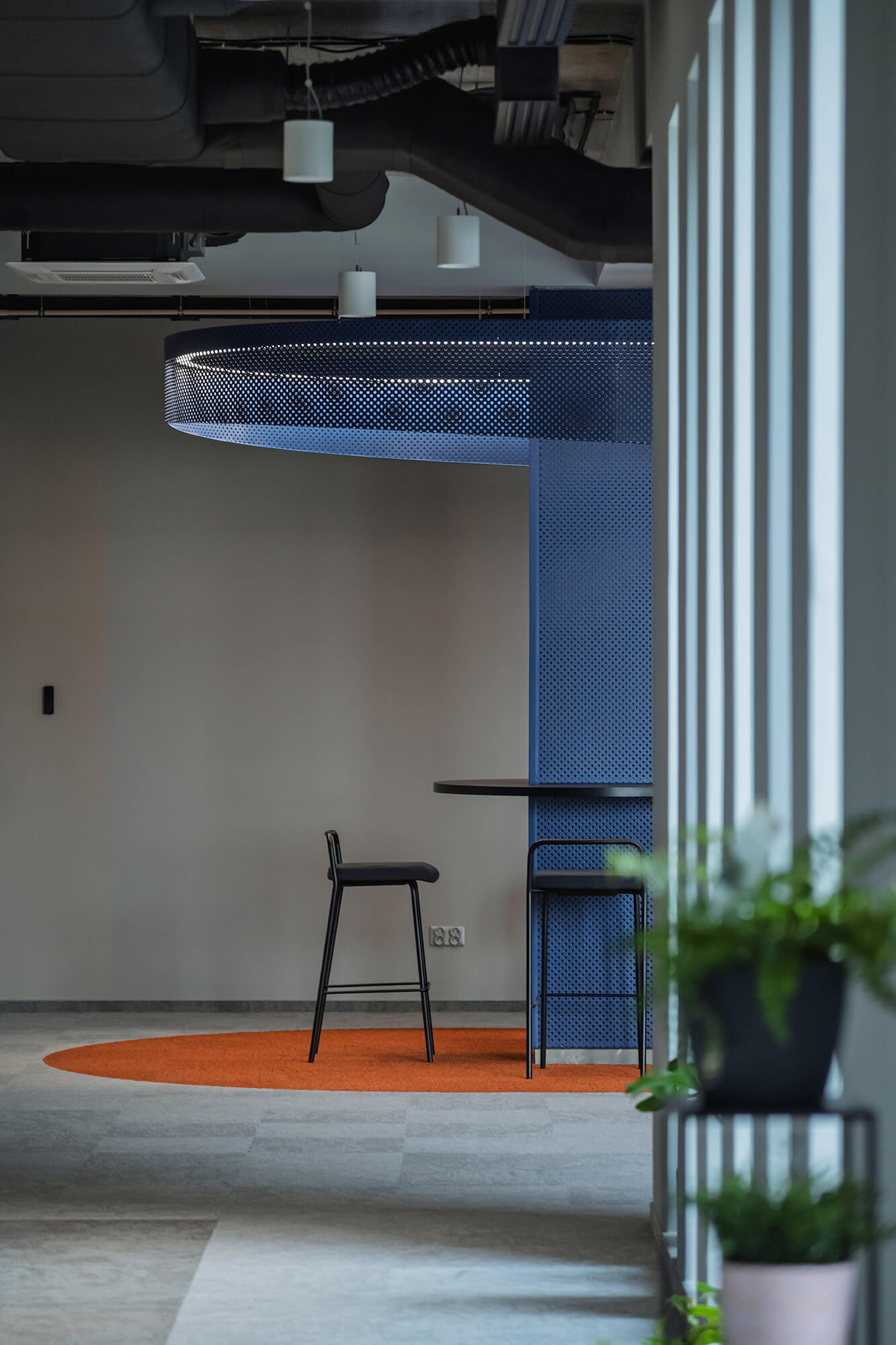 Nowoczesna przestrzeń biurowa z niebieskim panelem, stołkami barowymi i pomarańczowym dywanem.