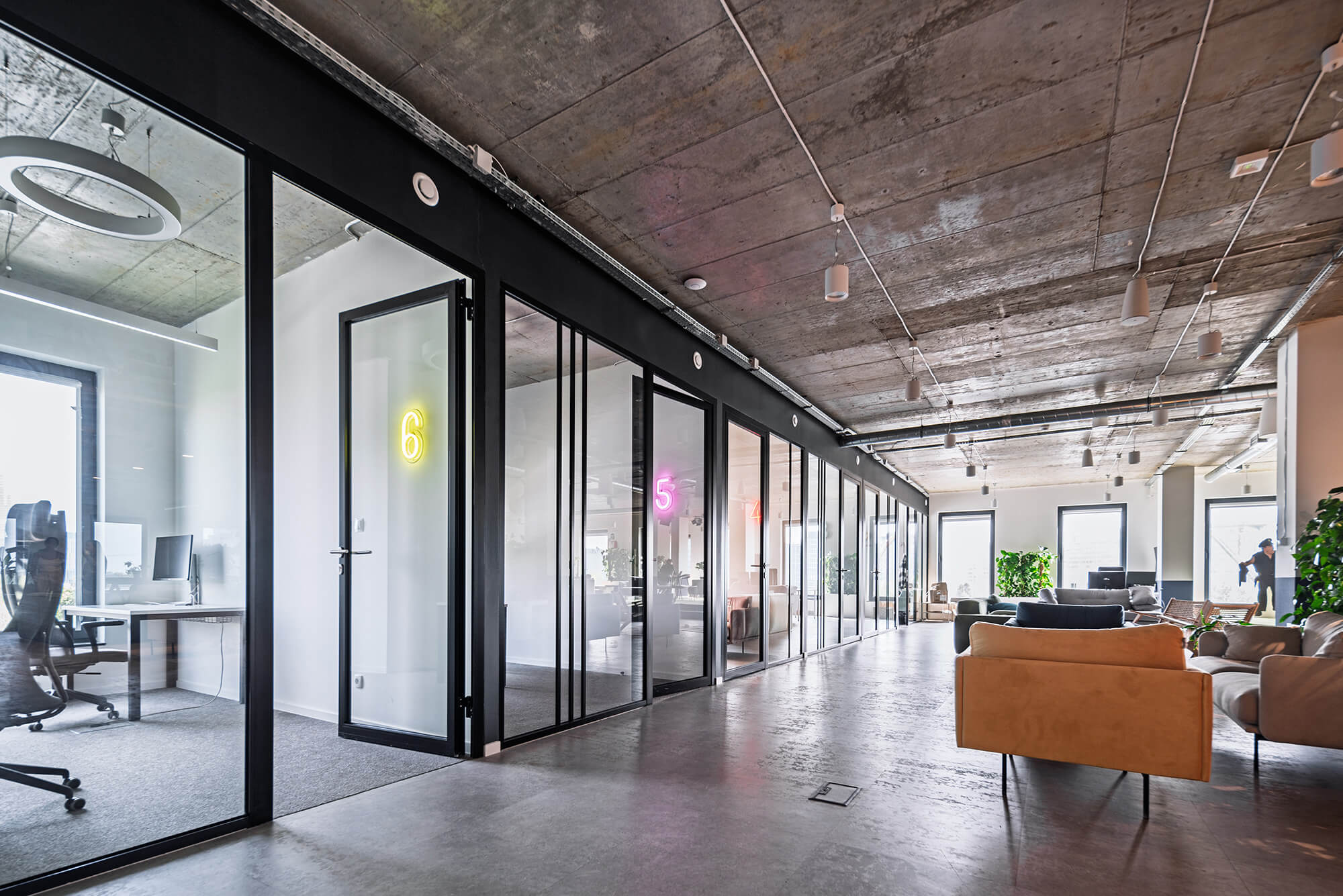 Nowoczesne biuro z betonowym sufitem, szklanymi ścianami i neonowymi numerami pokoi.