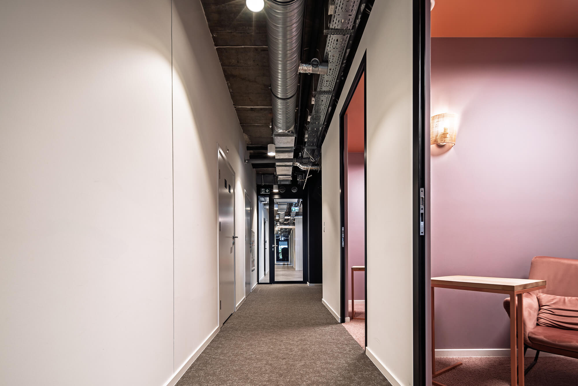 Korytarz biurowy z widocznymi instalacjami pod sufitem i wejściem do pokoju z fotelem.