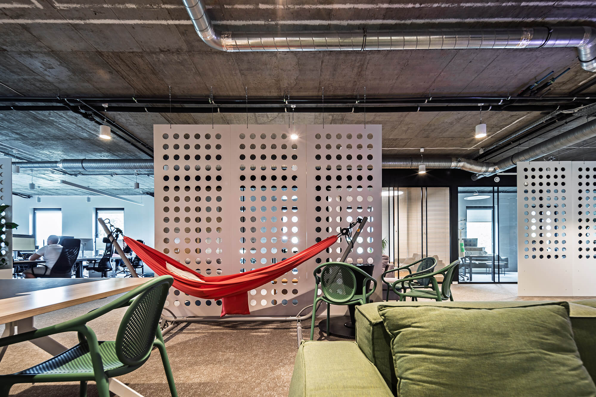 Nowoczesne biuro z czerwonym hamakiem, zielonymi krzesłami i surowym betonowym sufitem.